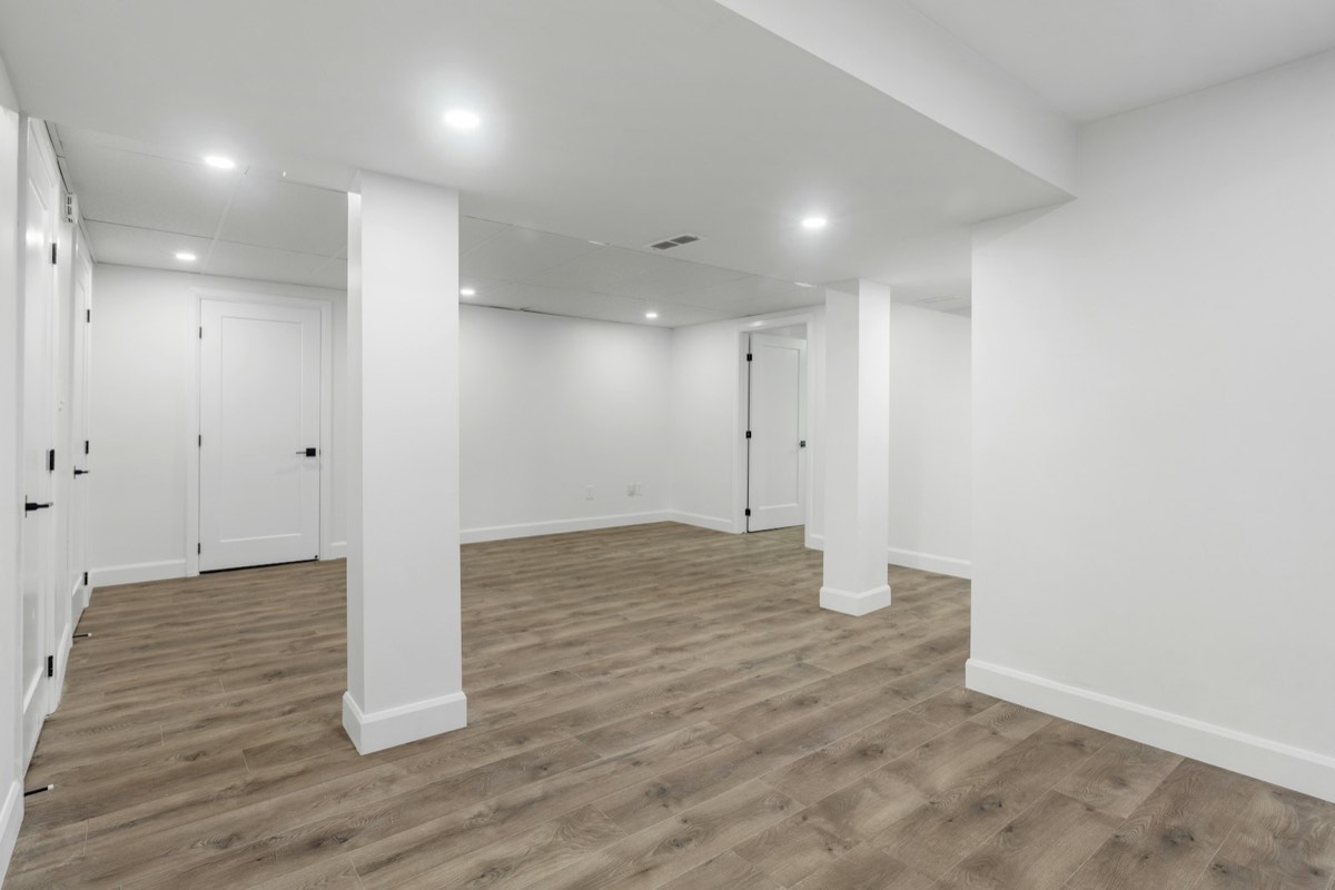 A finished basement with white painted walls, boxed support columns and wood-look plank flooring